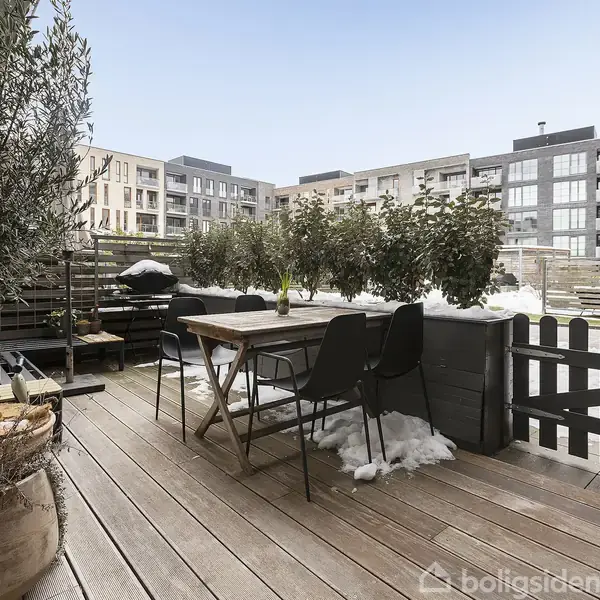 En træterrasse med bord og stole, omgivet af planter i krukker. Terrassen er omkranset af et lavt hegn og har udsigt til en moderne etageejendom i baggrunden. Snedækket jord ses omkring terrassen.