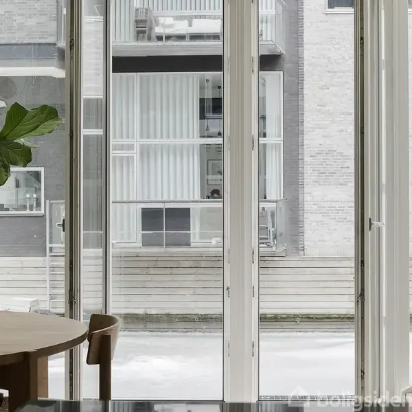 Et rundt spisebord med stole står tæt ved store glasdøre, der vender ud mod en moderne bygning udendørs. En stor potteplante til venstre giver et strejf af natur.