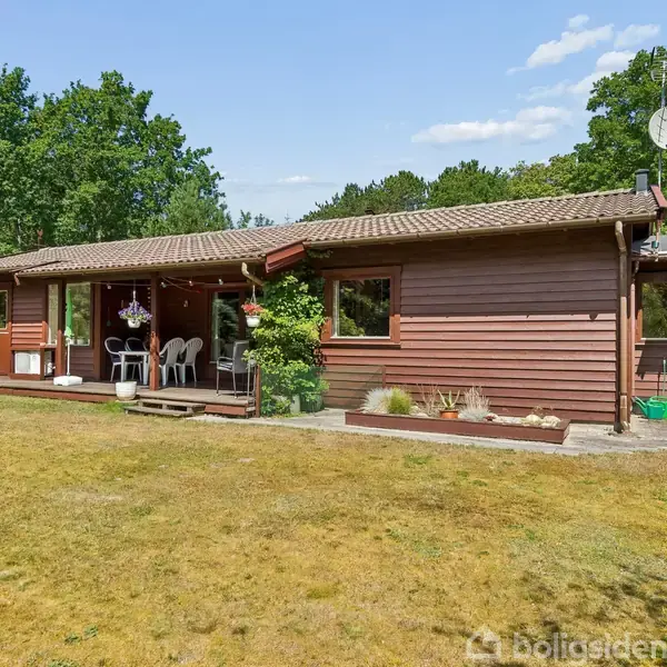En brun, træbeklædt bungalow med terrasse, omgivet af en græsplæne og skov. Havemøbler står på terrassen. Skyfri himmel og frodige grønne træer i baggrunden.