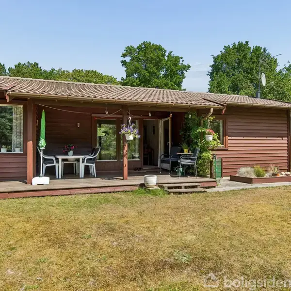 Træhus med veranda udstyret med havemøbler i en grøn have. Omgivet af græsplæne og træer, ses en grill og blomsterpotter.