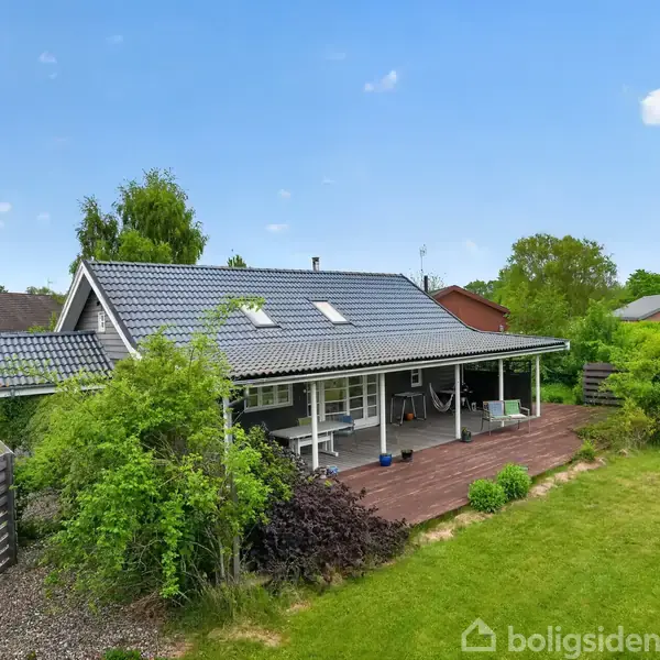 Et hus med gråt tag står omgivet af grønt løv. Det har en overdækket terrasse med borde og stole, og græsplænen strækker sig ud fra terrassen under en blå himmel.