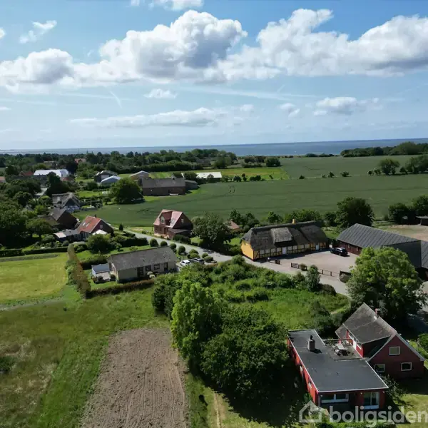 Landbrugsejendom spredt over et frodigt landskab med grønne marker og flere bygninger. Bygningerne er omgivet af marker og træer, og i horisonten kan man se en vandoverflade.