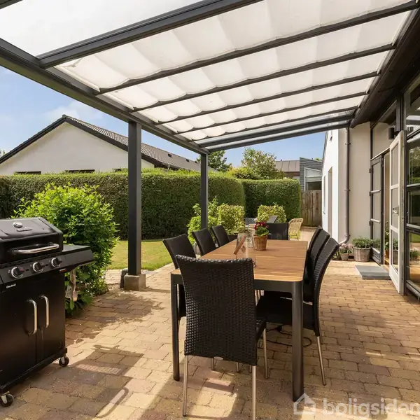 En overdækket terrasse med et langt bord og seks stole står ved siden af en grill. Haven er omgivet af grønne hække, og en moderne bygning ses i baggrunden.