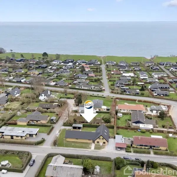 Hus markeret med EDC-logo ligger i et beboelsesområde fyldt med mange villaer. Husene er omgivet af grønne haver, og i baggrunden ses havet og grønne marker.