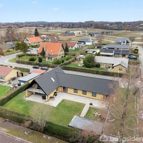 En lys murstenshus med mørkt tag ligger i et boligkvarter. Huset er omgivet af en hæk og græsplæne. Flere huse og veje ses i baggrunden under en klar himmel.