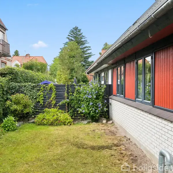 Et rødt og hvidt hus står langs en velholdt grøn græsplæne med omgivende hække og buske. En anden bygning kan ses til venstre. Himmel er klar og blå.