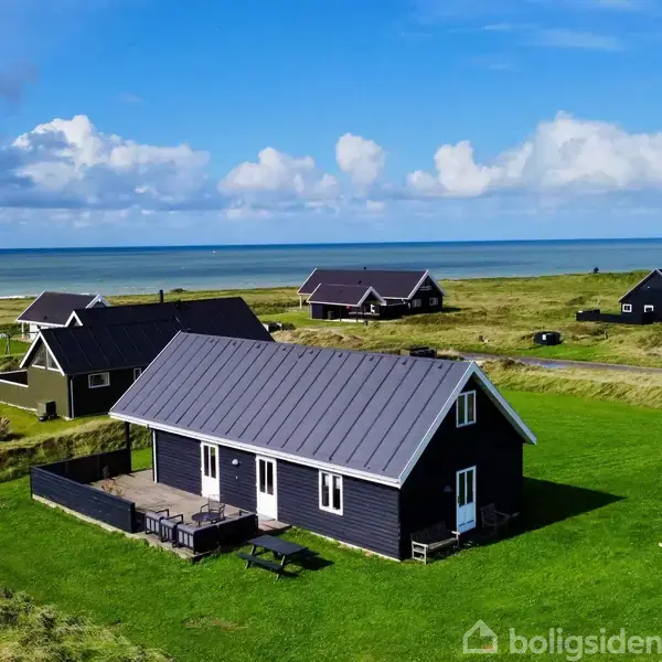 Sommerhus med sort facade og gråt tag står på en grøn græsplæne med havet i baggrunden. Flere huse er synlige i det omkringliggende landskab.