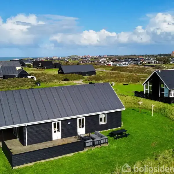 Et sort træhus står på en grøn græsplæne med veranda. Området består af flere huse og klitlandskab. I baggrunden ses en by med en kirke og kystlinjen.