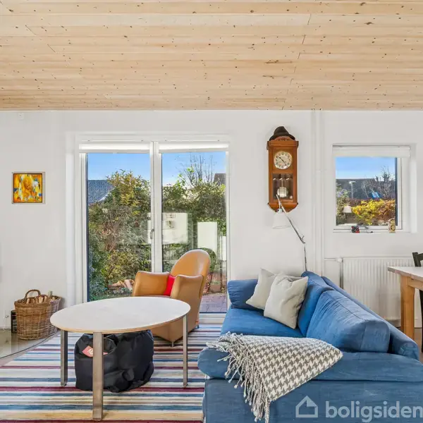 Brændeovn står i hjørnet og opvarmer stuen. Blå sofa og rund bord er midtpunkt, omringet af farverig stribet tæppe. Store vinduer giver udsigt til have og dagslys. Vægur hænger på væggen.
