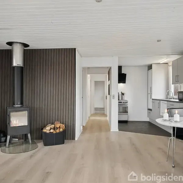 En moderne stue med en brændeovn ved en træpanelvæg. En kurv med brænde står ved siden af. I baggrunden ses et køkken med hvide skabe og et spisebord.