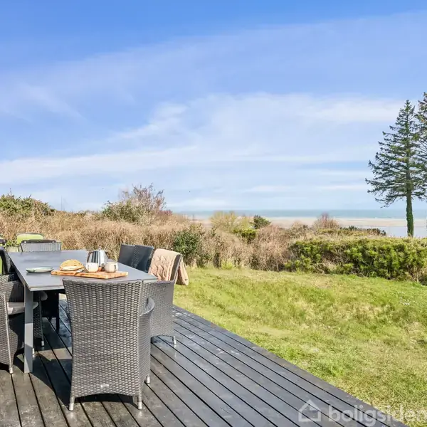Havebord med seks stole står på en træterrasse. Udsigt over græsplæne, klitter og havet i baggrunden under en blå himmel.