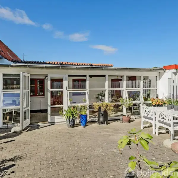 Terrasse omgivet af husfacader med rødt tag. Et hvidt havebord-sæt med stole står blandt mange planter i potter. En klar, blå himmel danner en lys og luftig atmosfære.