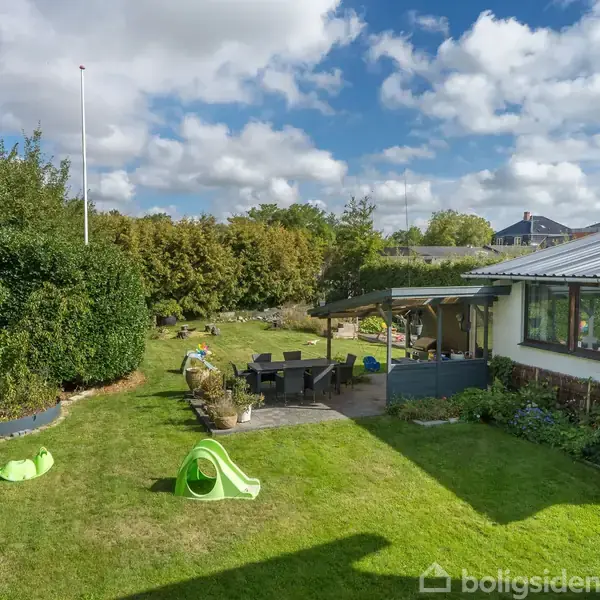 En grøn have med en lille rutsjebane og legeredskaber. En terrasse med spisebordssæt er placeret ved siden af et hvidt hus. Blå himmel med skyer i baggrunden.