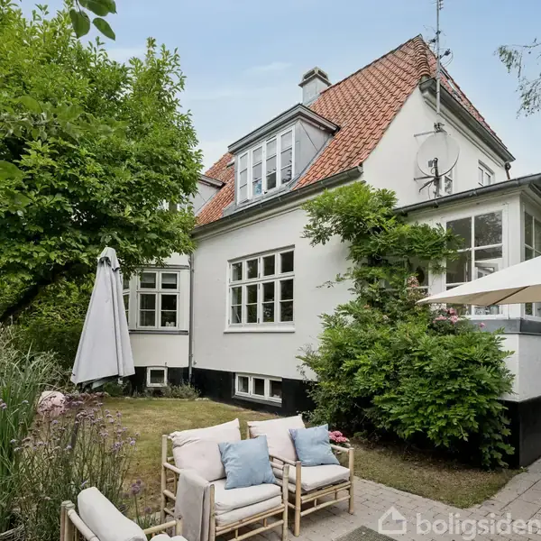 Huset står stille omgivet af grøn frodig have med træer og buske; foran er en terrasse indrettet med sofaer og parasol, hvilket skaber en hyggelig udendørs opholdsplads.
