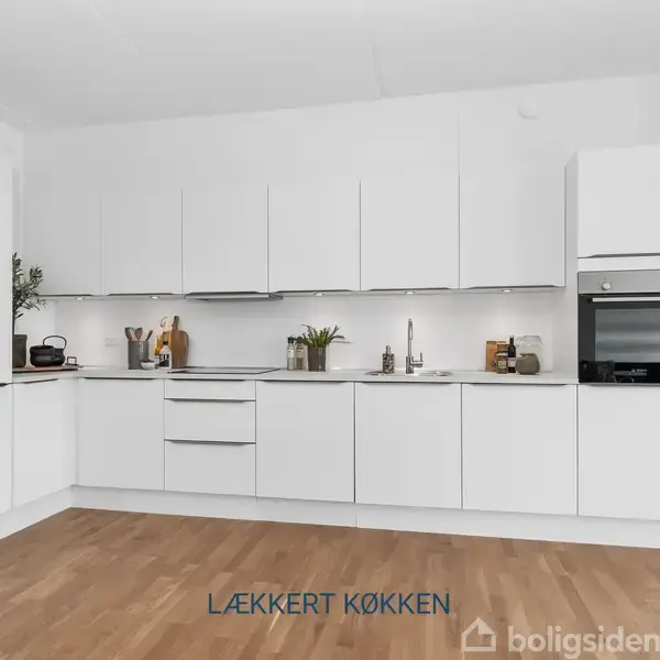 Et moderne køkken med hvide skabe og ovn indbygget i højre side. Planter og køkkenredskaber står på bordpladen. Teksten "LÆKKERT KØKKEN" vises på gulvet.
