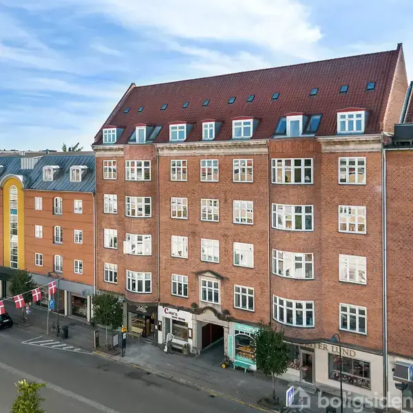 Bygning med rødt murstensværk og mansardtag, stående langs en gade med butikker i stueetagen. Der er hængt danske flag mellem træerne på fortovet.