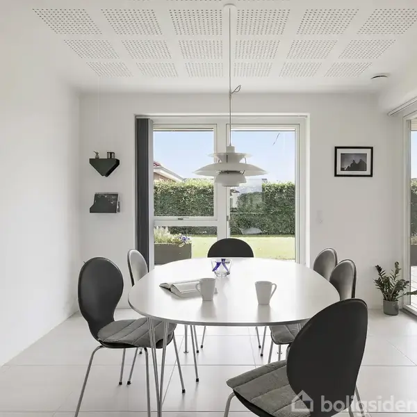 Et ovalt spisebord med seks stole står i et lyst rum. En moderne lampe hænger over bordet. Store vinduer viser en have med veltrimmede buske.