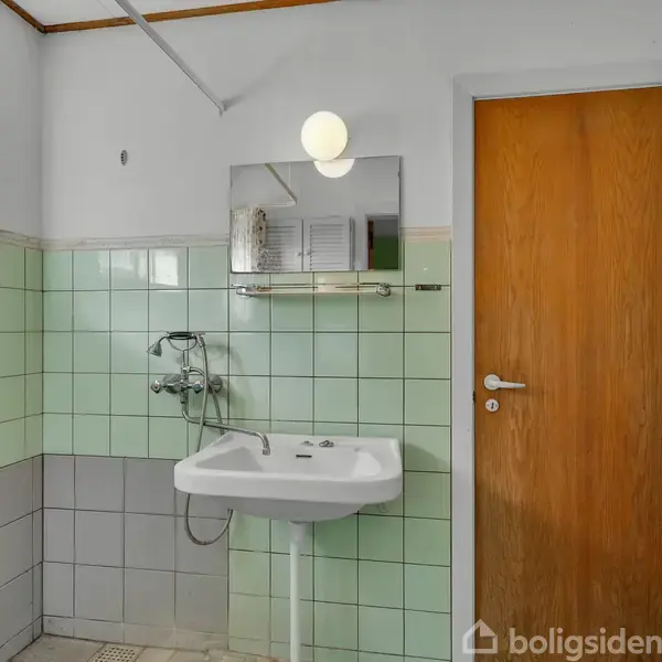 En hvid håndvask hænger på en væg med mintgrønne fliser, under et spejl med en rund lampe. Til højre ses en trædør og en radiator.