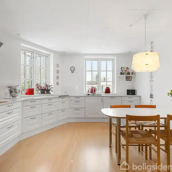 Køkken med hvide skabe og trælignende gulv; spisebord med fire stole; lysekrone hænger over bordet; udsigt til grøn beplantning; dekorationer og ur på væg; sollys gennem vinduerne.