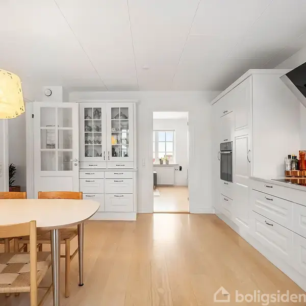 En lyst køkken med et rundt spisebord og fire stole, hvor der hænger en lampe over bordet. Køkkenet har hvide skabe, indbygget ovn og farverig flisevæg bag kogepladen.