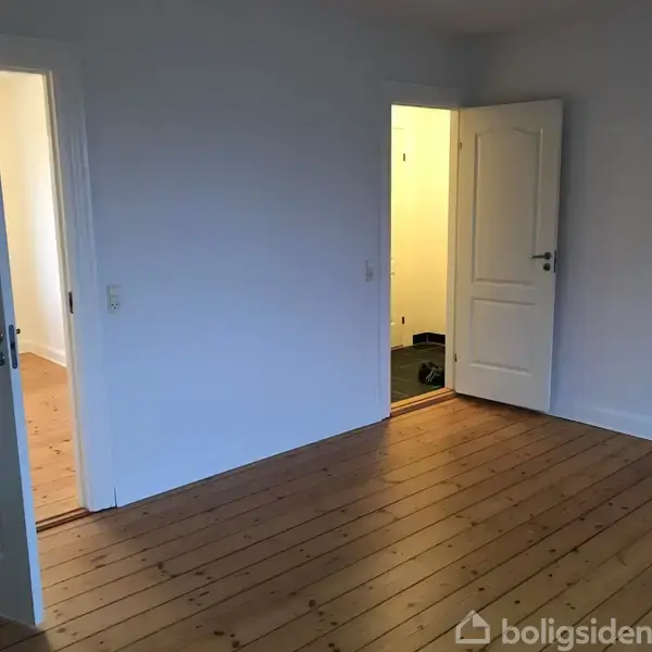 Et tomt værelse med trægulve åbner ind i en lys gang. Døre står åbne, og lyset fra gangen skaber kontrast.