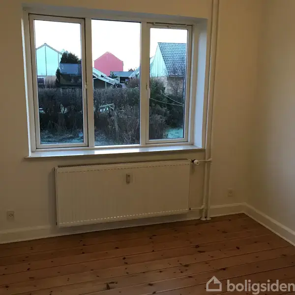 Et tomt værelse med trægulv har et vindue og radiator. Udenfor ses en have med buske og huse i baggrunden. Væggene er lyse.