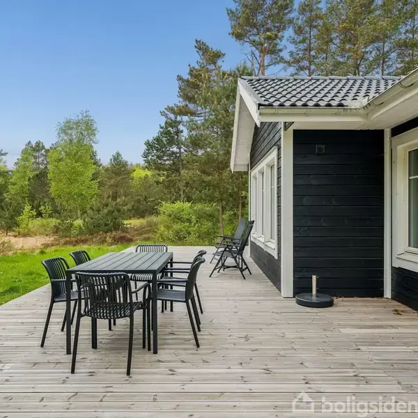 Et sort træhus med en stor træterrasse står i skovomgivelser. Terrassen har et bord og flere stole, og omgivet af grønne træer og en græsplæne i baggrunden.