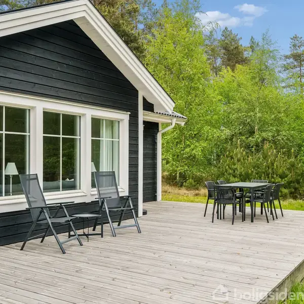 Et sort træhus med store vinduer. To sorte stole står på den træbelagte terrasse, hvor der også er et bord med fire stole. Omgivet af grønne træer.