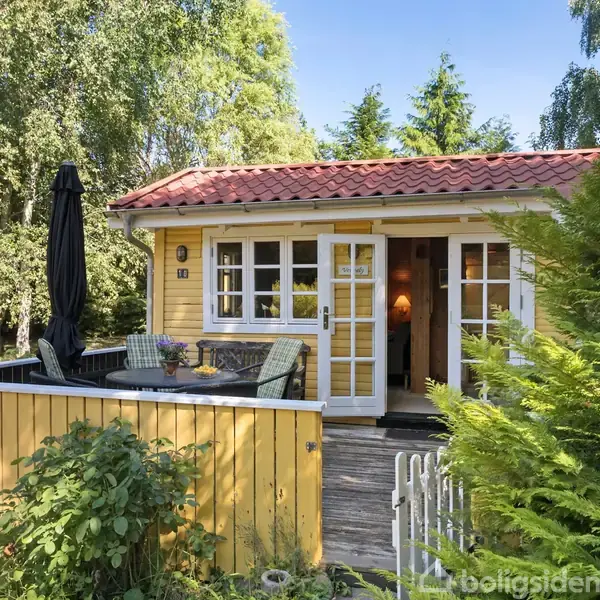Et gult sommerhus med terrasse ligger omgivet af frodig vegetation. Terrassen har et bord, stole og en opslået parasol. Huset har rødt tegltag og hvidrammede vinduer.
