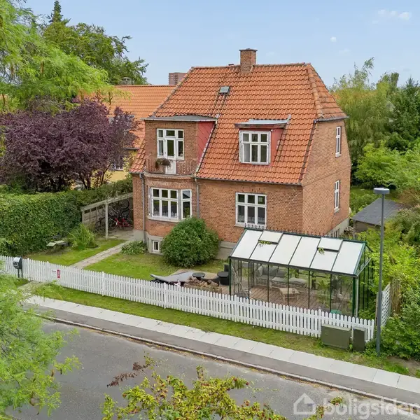 Et rødt murstenshus med rødt tegltag står ved en grøn have, omringet af hvide stakitter og en stor glasudestue. Huset ligger i et roligt villakvarter.