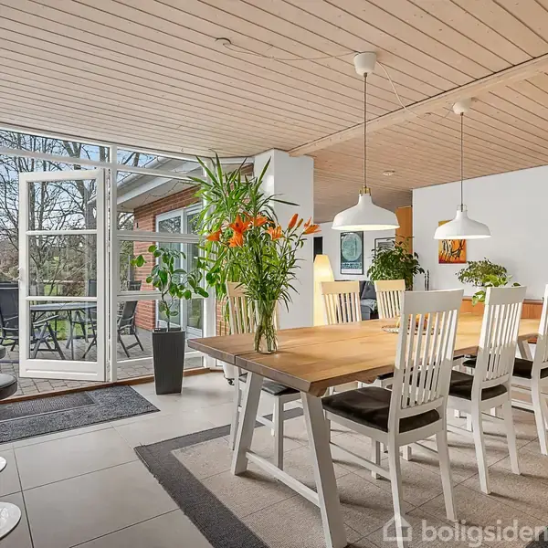 Et spisebord med otte hvide stole står i et lyst rum med store vinduer. Friske blomster i vase pynter bordet. Udenfor ses en terrasse med et bord og stole.