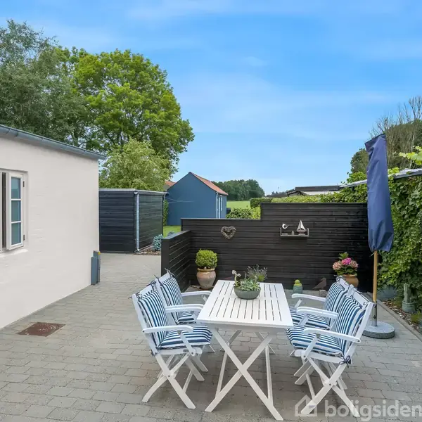 Havebord med seks stole står på flisebelagt terrasse ved siden af hvid bygning. Omkring ses grønne planter og blåmalet træhegn med dekorative elementer.