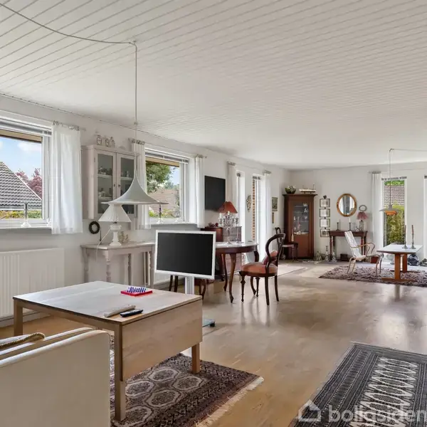 Stue med lyse vægge og trægulv, indrettet med sofa, sofabord og TV. Store vinduer slipper naturligt lys ind. Møbler inkluderer spisebord og skabe. Tæppe på gulvet.