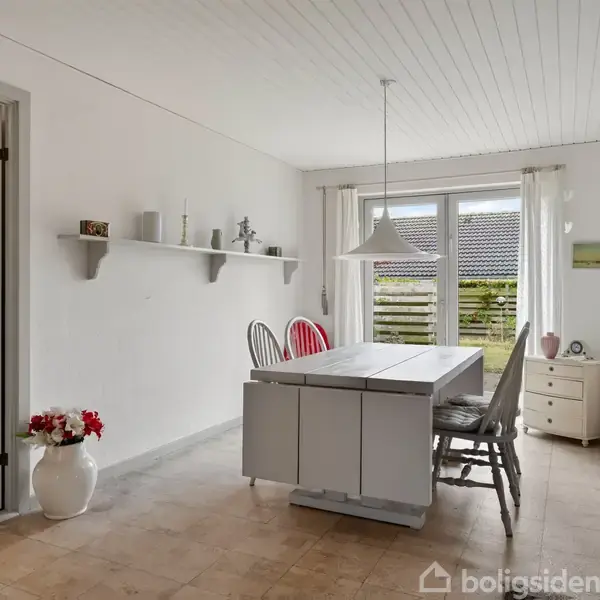 Spisebord med stole står i et lyst rum med lyse vægge og klinkegulv. En hylde med dekorationer hænger på væggen. Store vinduer giver udsigt til terrasse og have.