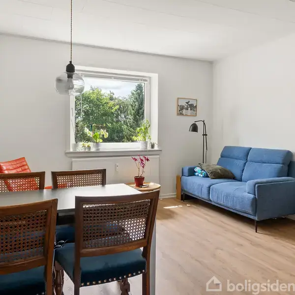 Et spisebord med stole står ved et vindue. En blå sofa står til højre med en gulvlampe og en plante. Værelset har lyse vægge og trægulv.