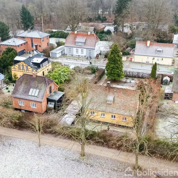 Række af små huse står stille langs en træbevokset vej i et vinterpræget boligområde. Sne dækker jorden, mens baggrunden domineres af nøgne træer og flere huse.