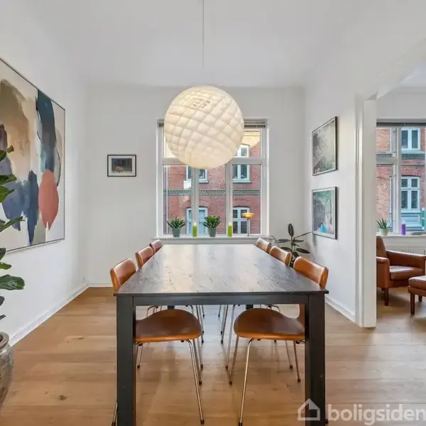 Et spisebord med otte stole står i et lyst rum. En stor plante står ved siden af bordet, og en rund lampe hænger over bordet. Vinduet viser en udsigt til nabo-bygninger. Tekst: "indretningsforslag".