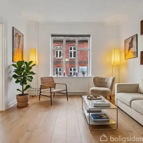 Stue med sofa, lænestol, og sofabord i et lyst rum. Store vinduer viser røde murstensbygninger udenfor. En plante og lamper tilføjer dekorative elementer. I baggrunden ses en åbning til et spiseområde.
