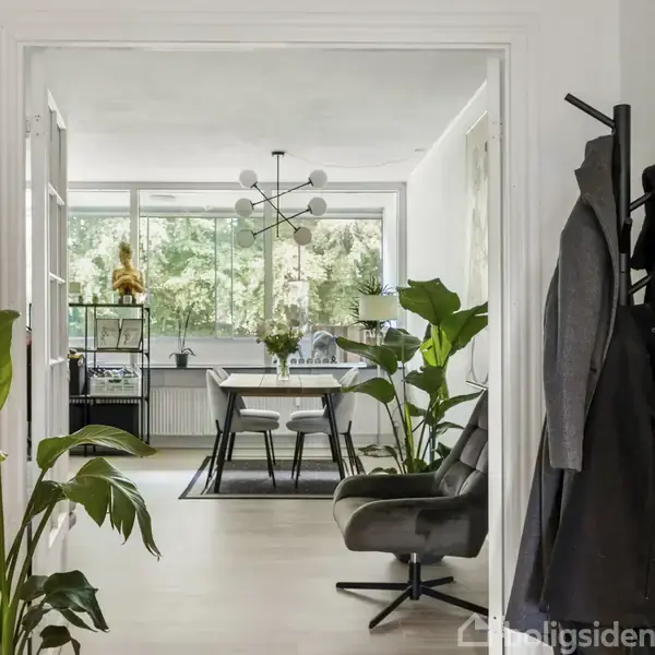 Spisestue med et bord og fire stole står placeret under en moderne, geometrisk loftlampe. Vinduerne i baggrunden giver udsigt til træer udenfor. Grønne planter og lyse vægge omringer rummet.