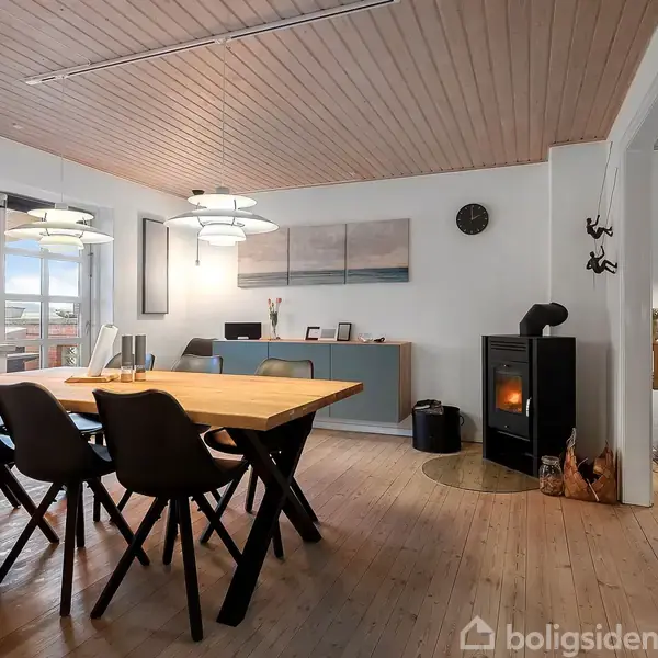 Spisebord med seks stole står i et lyst rum med trægulv og træloft. En brændeovn står i hjørnet, og store vinduer giver udsigt til en terrasse.