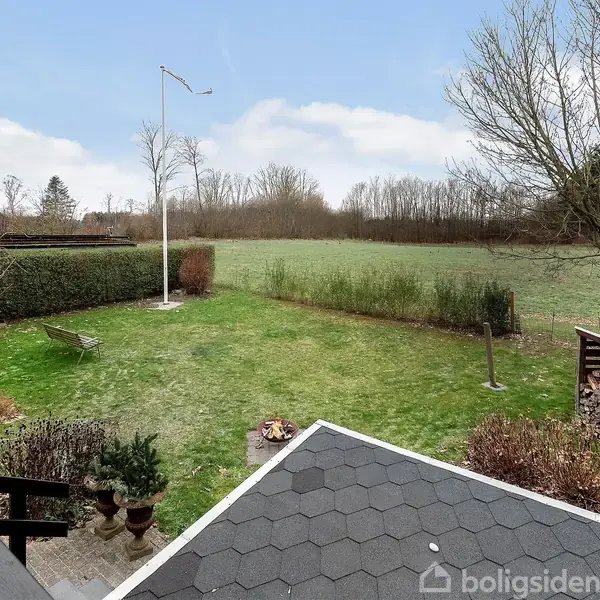 Græsplæne strækker sig ud fra en terrasse med sort tag, omgivet af træer og buske. Et hegn markerer grænsen til et større grønt område.