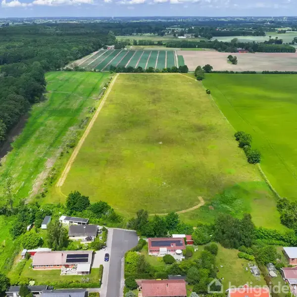 En lang græsmark strækker sig gennem landbrugsområder, omgivet af grønne marker og træer, mens nogle huse ligger i forgrunden langs en vej.