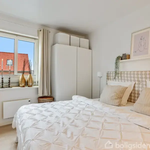 Seng med hvid dyne står i et lyst soveværelse. Gardiner omkranser et vindue med udsigt til røde tegltage. Dekorationer og planter er placeret på hylder over sengen.