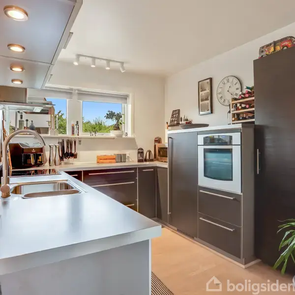 Køkken i moderne stil med overflader af rustfrit stål og sort kabinet. En indbygget ovn står til højre, mens planter og vægdekorationer skaber en indbydende atmosfære.