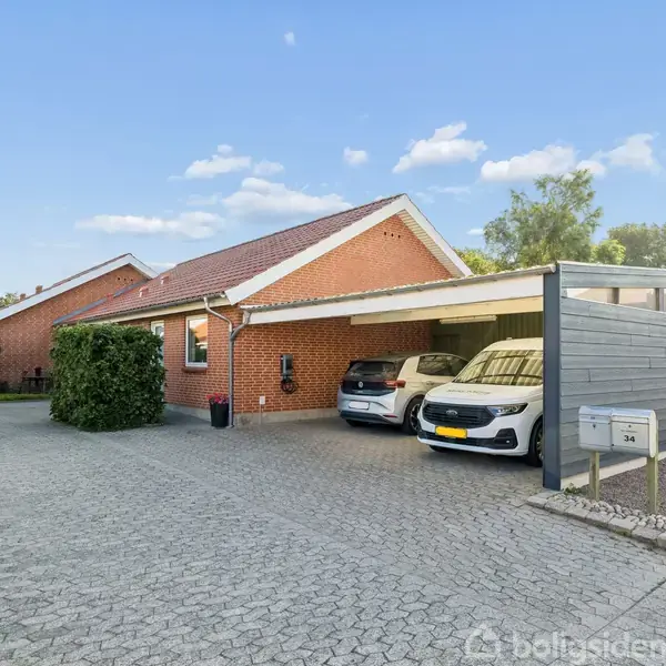Rød murstenshus med skråt tag og tilhørende carport, hvor to biler holder parkeret. Solskinsdag med blå himmel og få skyer omgivet af grønne buske og træer. Sti fører til indgang.