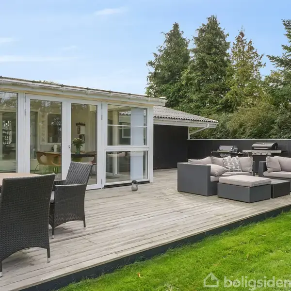 En lys træterrasse med havemøbler og grill omgivet af grøn have og træer, set fra siden af et moderne hus med store glasvinduer.