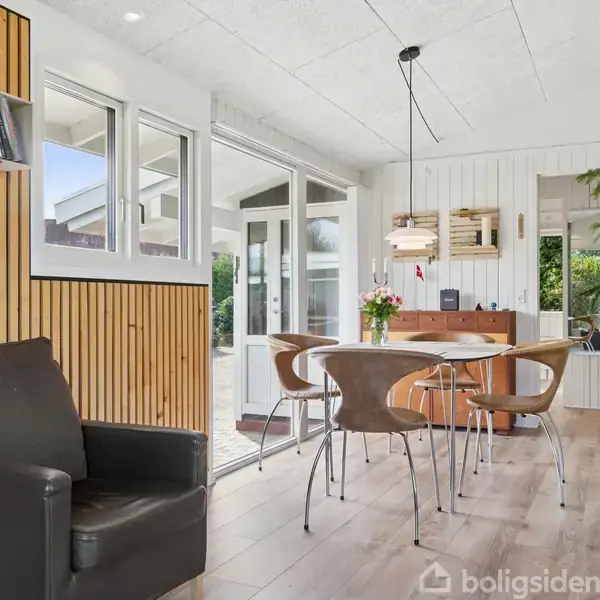 En moderne spisestue med runde bord og stole står i et lyst rum med trægulve. Et skab med planter og udsigt til grøn have gennem store vinduer.