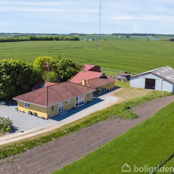 En gul villa med rødt tag står blandt træer, ved siden af en stor lade. Bygningerne er omgivet af vidtstrakte grønne marker under en blå himmel.