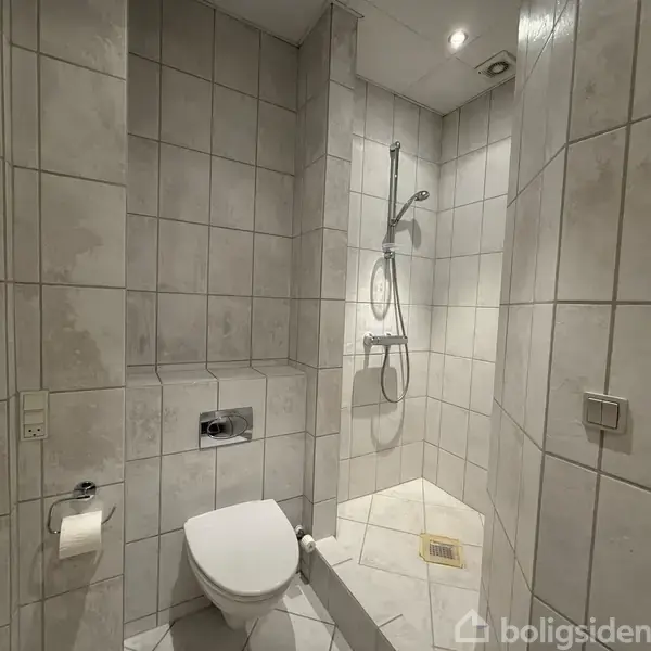 Badeværelse med hvidt flisegulv og vægge har et hvidt skab med vask og spejl. Begrænset opbevaring under vasken. Hvid dør ved væggen. Toiletrulle på væggen.