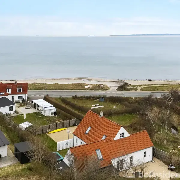 Hus med rødt tag står blandt andre boliger nær kysten. Baggrunden viser havet og en åben himmel. En vej adskiller husene fra stranden.
